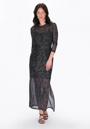 Kvinde iført en gennemsigtig sort kjole med lange ærmer og blomsterprint samt sorte sandaler med stropper, stående foran en ensfarvet hvid baggrund.