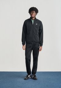 Μαύρο αθλητικό σετ Nike που περιλαμβάνει ζακέτα με φερμουάρ και αντιστοιχούντα παντελόνια. Κατασκευασμένο από λείο ύφασμα με ελαστικές λεπτομέρειες. Το μοντέλο φοράει μαύρα αθλητικά παπούτσια.