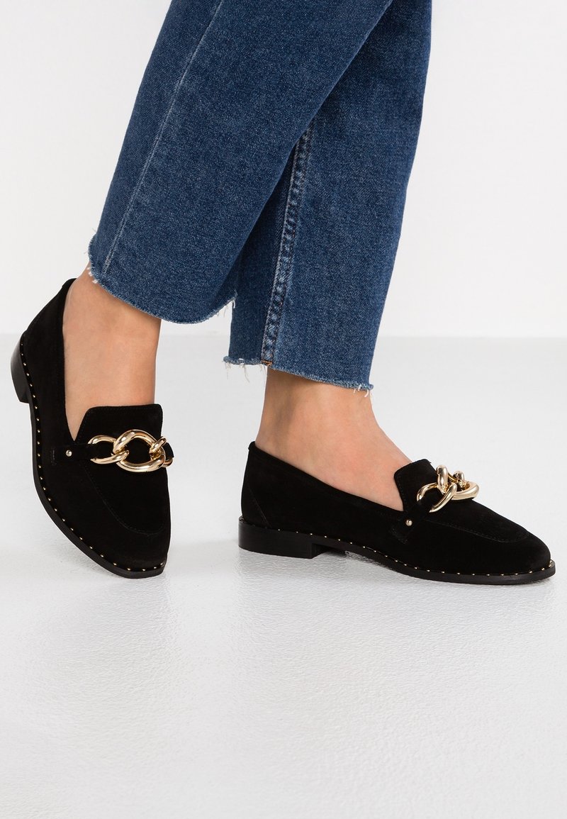 Mocassins en daim noir avec un accent de chaîne dorée, semelle plate et détails cloutés le long du bord ; associés à un jean court en denim.