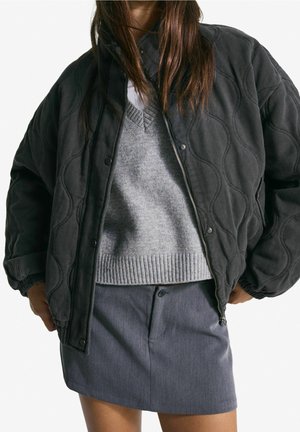 Kvinde iført en sort quiltet jakke over en grå sweater og en mørkegrå knap-nederdel, med hænderne i jakkelommerne.