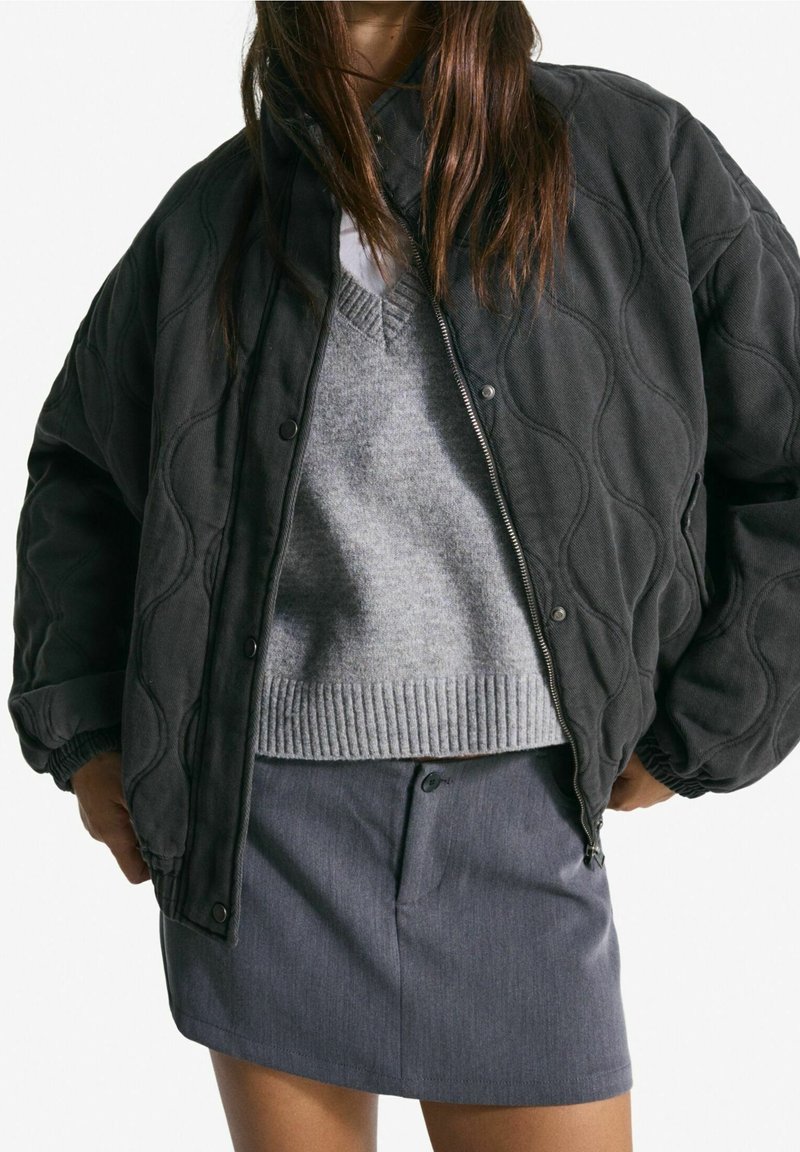 Femme portant une veste matelassée noire par-dessus un pull gris et une jupe boutonnée gris foncé, les mains dans les poches de la veste.