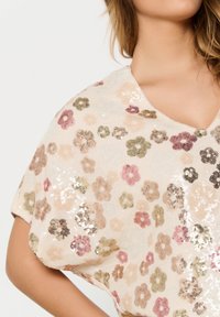 Donna che indossa un top beige con paillettes e motivi floreali multicolore, mostrando il décolleté e la parte superiore del braccio su uno sfondo bianco.