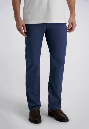 Mann trägt marineblaue Chinos, braune Lederloafer und ein weißes Hemd, steht vor einem einfarbigen Hintergrund.