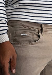 Pantalones de mezclilla beige con un bolsillo delantero, que presentan una etiqueta plateada de "Petrolind". Suéter a rayas en colores azul marino y blanco que cubre el brazo.