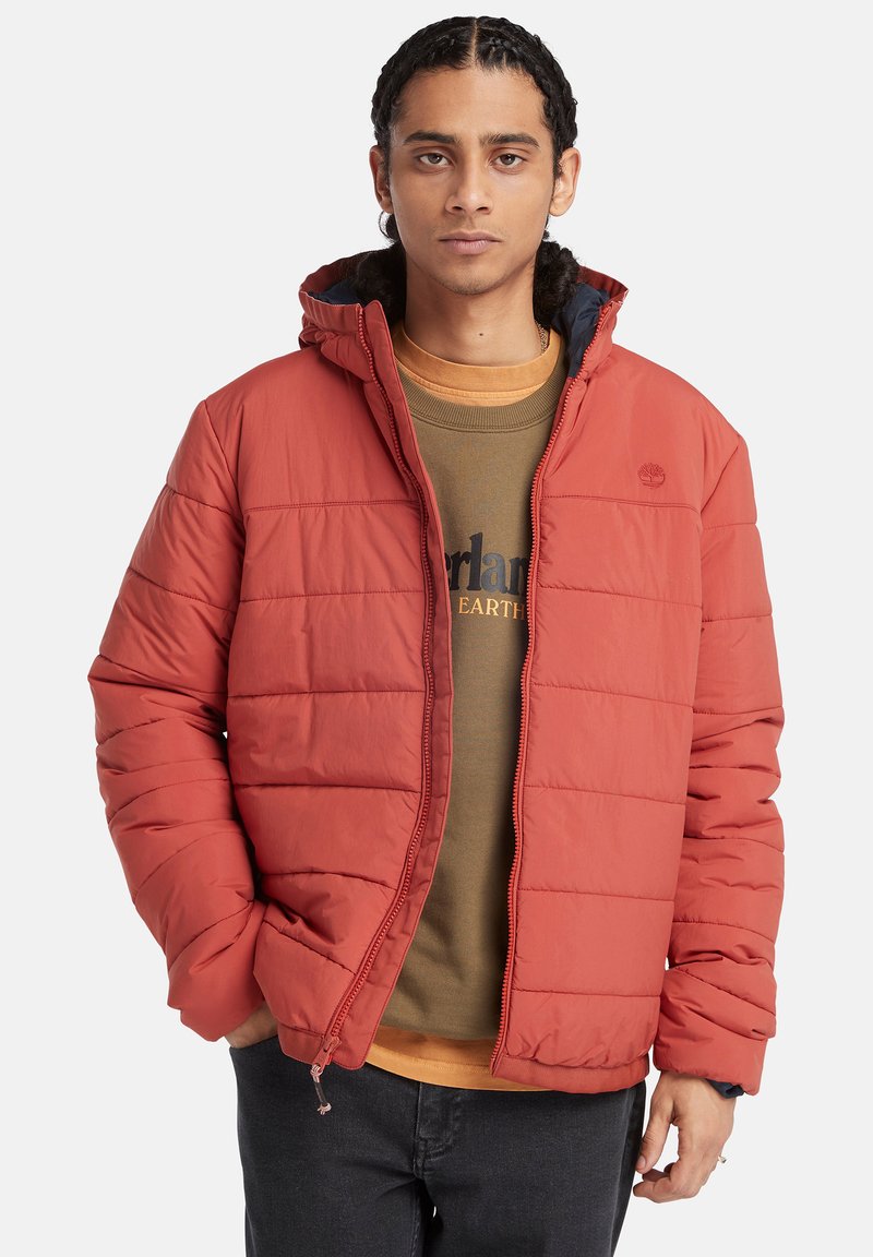 Rote Daunenjacke mit Kapuze, im gesteppten Design, mit Reißverschluß und elastischen Bündchen. Getragen über einem braunen Sweatshirt.