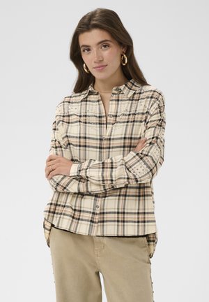 Giovane donna con lunghi capelli castani che indossa una camicia beige a quadri con borchie e pantaloni beige, braccia incrociate, espressione neutra su sfondo bianco.