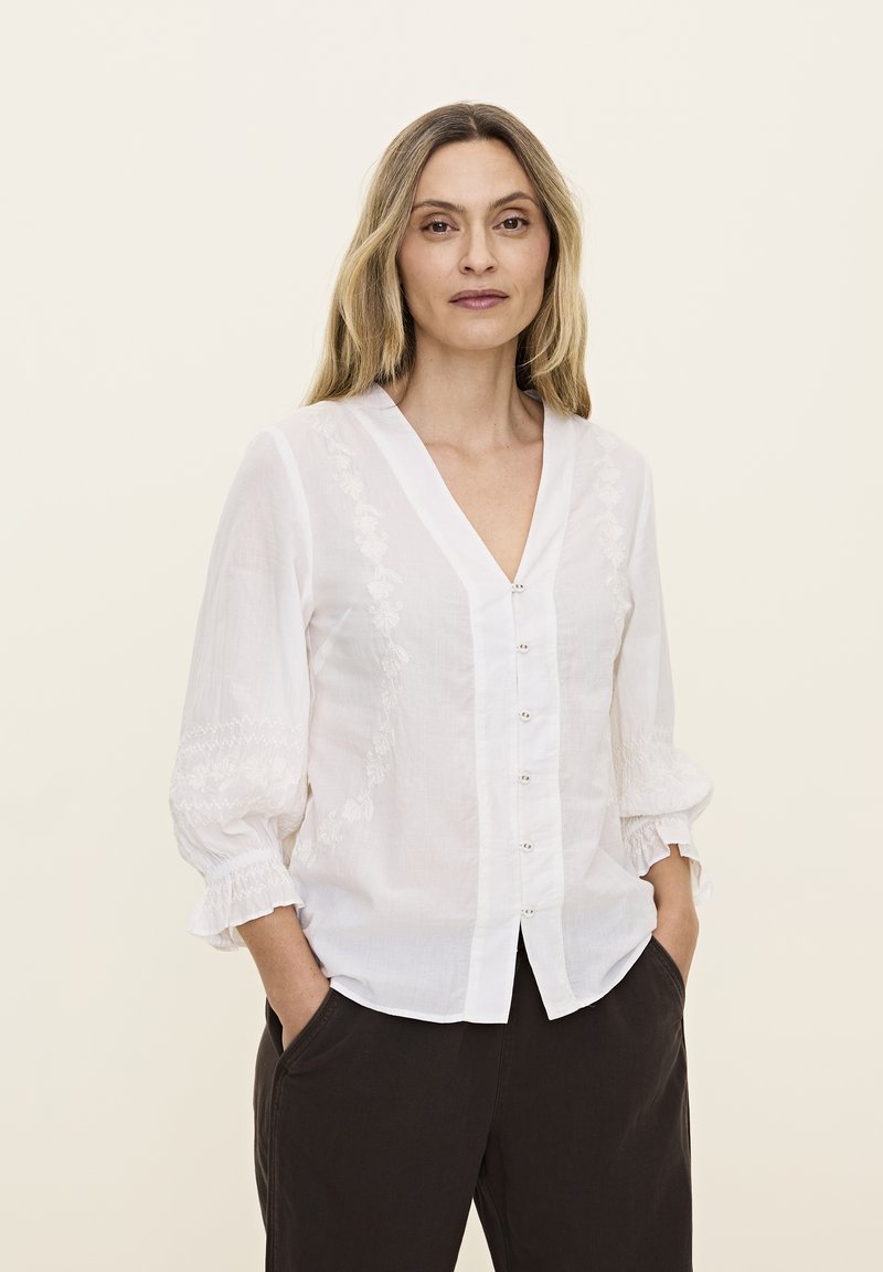 Blouse blanche à boutons avec un décolleté en V, ornée de détails floraux brodés sur les côtés et de manches froncées. Associée à un pantalon sombre.
