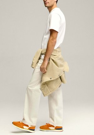 Witte katoenen T-shirt, beige jas om de taille geknoopt, crèmekleurige broek en oranje suède sneakers met blauwe en witte accenten.