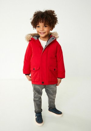 Veste à capuche rouge avec une bordure en fausse fourrure, deux poches avant et une doublure grise. Associée à un pantalon gris et des baskets bleues.