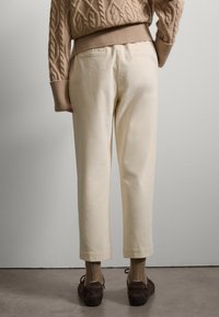 Suéter de punto beige sobre pantalones rectos de color crema con una textura suave, con tobillos ajustados y zapatos de ante marrón.