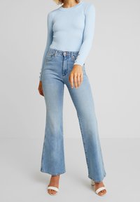 Kvinna som bär en ljusblå ribbad långärmad topp, högmidjade ljusblå utsvängda jeans och vita öppetåiga klacksandaler.