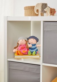 Zwei Plüschpuppen sitzen auf einem Buch, eine in einem rosa Kleid mit orangefarbenen Akzenten und einem grauen Hut, die andere in einem blau gestreiften Outfit und einem dunklen Hut.