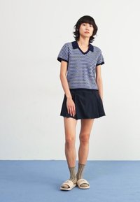 Polo en tricot bleu marine et bleu clair à pois, avec un col, associé à une jupe foncée, des chaussettes vert olive et des sandales à enfiler beige.