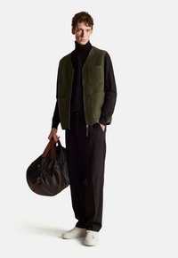 Olivgrüner Fleece-Westen mit Fronttaschen, kombiniert mit einem schwarzen Rollkragenpullover, schwarzen Hosen und weißen Turnschuhen; haltend eine schwarze Sporttasche.