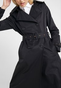 Svart trenchcoat med dubbelknäppta knappar, bälte i midjan och stora slag. Tillverkad av ett mjukt, lätt material.