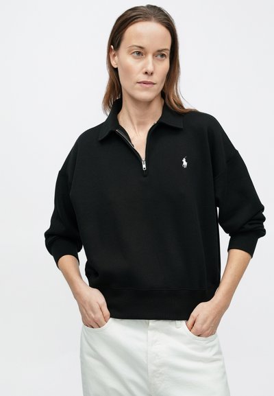 Svart genser med halvglidelås og ribbet kant nederst. Har en liten hvit logo på brystet. Mykt stoff med en avslappet passform.