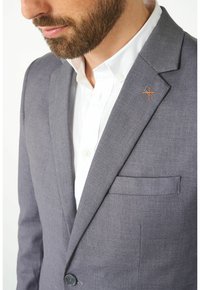 Grauer Blazer aus strukturiertem Stoff, mit einem einzigen Knopfverschluss, Reverskragen und einer dezenten orange-grauen Reversnadel. Darunter ein weißes Hemd.