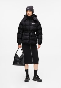 Schwarze Puffjacke mit einem taillierten Gürtel, gerippten Bündchen und Logo. Getragen über einem schwarzen knielangen Rock, kombiniert mit schwarzen Sneakers und einer Tasche.