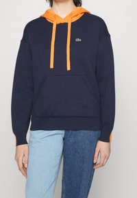 Navy hoodie with orange drawstrings and accents. Features a front pocket and ribbed cuffs. Cotton blend fabric, relaxed fit.