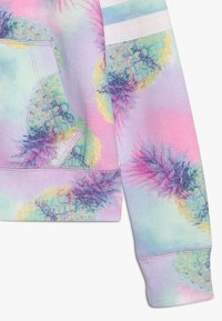 Hoodie pastel avec motif d'ananas en violet et jaune. Tissu doux avec une poche avant et un détail à rayures blanches sur la manche.