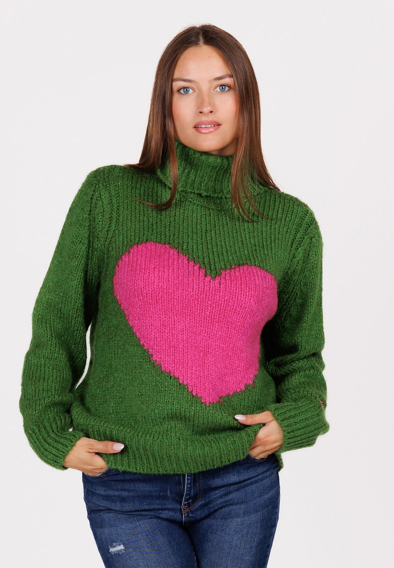 Vrouw met lang bruin haar, gekleed in een groene gebreide coltrui met een groot roze hart aan de voorkant, met handen in de zakken, en blauwe spijkerbroeken.