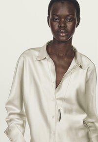 Camicia in satin color crema con colletto rilassato; presenta un profondo scollo ed è abbinata a una collana con pendente in argento.