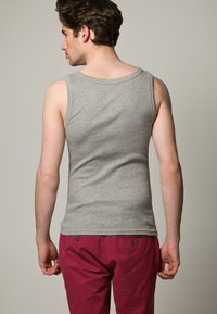 Hallikas puuvillane tank top ümmarguse kaelusega, ribakujuline tekstuur ja ilma varrukateta disain, kantud punaste pükstega. Fookus tagapoolses vaates.