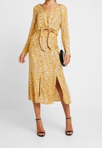 Kvinna i en gul leopardmönstrad långärmad klänning med framslitsar, svarta ankelremmar och håller en liten svart väska med kedja.