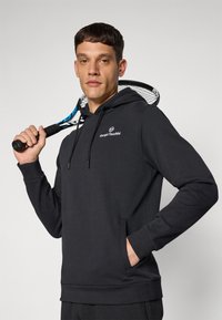 Svart hettegenser med snorer, frontlomme og logo. Glatt tekstur, atletisk passform, brukt av en person som holder en tennisracket.