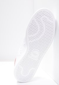 Biała podeszwa sportowa z heksagonalnym wzorem. Posiada gładką teksturę oraz czerwony akcent z boku. Logo wytłoczone w pobliżu pięty.