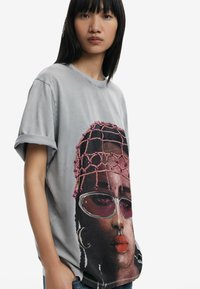 Sivá oversized tričko s grafickým portrétom v živých farbách, vrátane fialových akcentov a slnečných okuliarov, s rolovanými rukávmi.