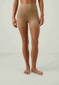 Sieviešu modele, kas valkā beige augstās vidukļa bezmugurkaula veidojošās šortus un atbilstošas atvērtās pirkstu kāju segas uz vienkārša gaiša fona.