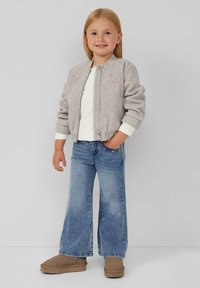 Graue Bomberjacke mit floralem Stickerei, weißes Hemd, ausgestellte blaue Jeans und braune Stiefel. Kind steht in einer entspannten Pose.