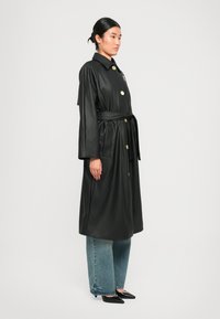 Cappotto lungo in finta pelle nera con colletto, bottoni dorati e cintura annodata; abbinato a jeans blu a gamba larga e scarpe nere a punta.