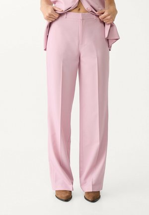 Vrouw draagt hoge taille, wijde roze broek gecombineerd met bruine puntige schoenen, houdt een bijpassend roze jasje open.