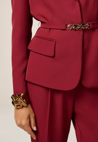 Costume bordeaux ajusté avec une coupe slim, comprenant un bracelet épais en or métallique avec un détail en nœud et une poche latérale sur la veste.