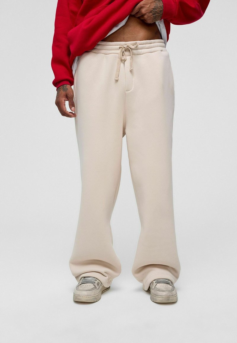 Beige joggers met een relaxed fit, voorzien van een elastische tailleband en een trekkoord. Het materiaal lijkt zacht en net textuur te hebben. Gepaard met grijze sneakers.