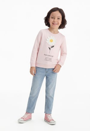 Enfant souriant aux cheveux bruns courts portant un sweat-shirt rose avec un motif de marguerite, un jean bleu clair et des baskets montantes roses.