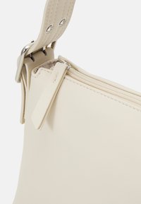 Beige synthetische Schultertasche mit glatter Textur, ausgestattet mit einem Reißverschluss und einem verstellbaren Riemen mit dekorativen Nähten und silbernem Beschlag.