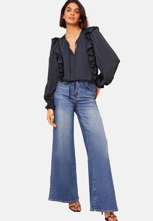Sort bluse med rynket design, v-hals og puffede ærmer, parret med høj talje vide blå jeans. Model står på en hvid baggrund.