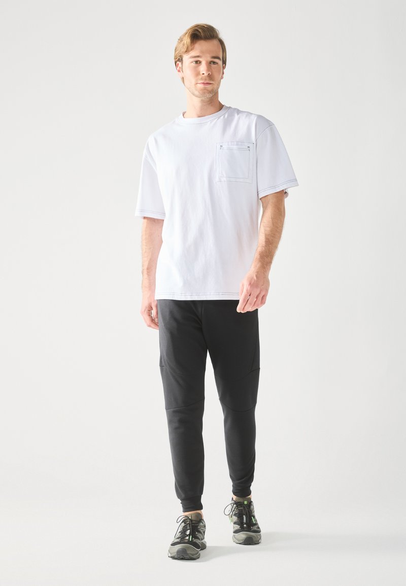 Valkoinen lyhythihainen T-paita, jossa on tasku, yhdistetty mustiin kapeisiin housuihin, joissa on sivutaskut ja saumat. Asusteena harmaat urheilukengät.