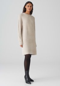 Vestito a maglione in beige chiaro, con maniche lunghe e scollatura rotonda. Indossato sopra collant neri con stivaletti neri a punta.