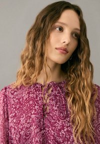 Lila Bluse mit Paisley-Muster, Schlaufenverschluss am Hals und langen Ärmeln, verziert mit floralen Akzenten und aus einem weichen, leichten Stoff.