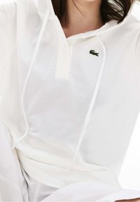 Vit huvtröja med pullovermodell i lätt mesh-tyg, med halv-zip krage och en liten grön Lacoste-logga på bröstet.