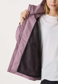 Person wearing white shirt shows interior mesh lining of open mauve jacket, focusing on jacket’s texture and pocket detail.