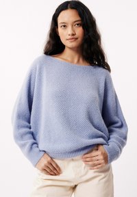 Pull en tricot bleu clair à coupe ample, décolleté bateau large et motif texturé. Manches longues avec poignets côtelés, porté sur un pantalon crème.