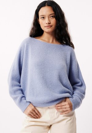 Pull en tricot bleu clair à coupe ample, décolleté bateau large et motif texturé. Manches longues avec poignets côtelés, porté sur un pantalon crème.