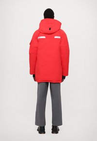 Rote isolierte Parka mit großer Kapuze, ausgestattet mit reflektierenden Streifen auf dem Rücken und schwarzen Akzenten. Kombiniert mit grau weitem Hosen und schwarzen Schuhen.