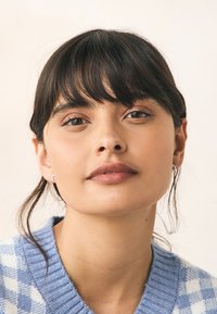 Plava i bijela džemper s kockastim uzorkom i rebrastim ovratnikom. Suptilne srebrne naušnice. Glatka koža i ravna, tamna kosa s resama.