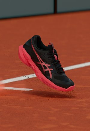 Musta ja punainen urheilukenkä potkaisee punaista savea tenniskentällä valkoisen viivan lähellä.